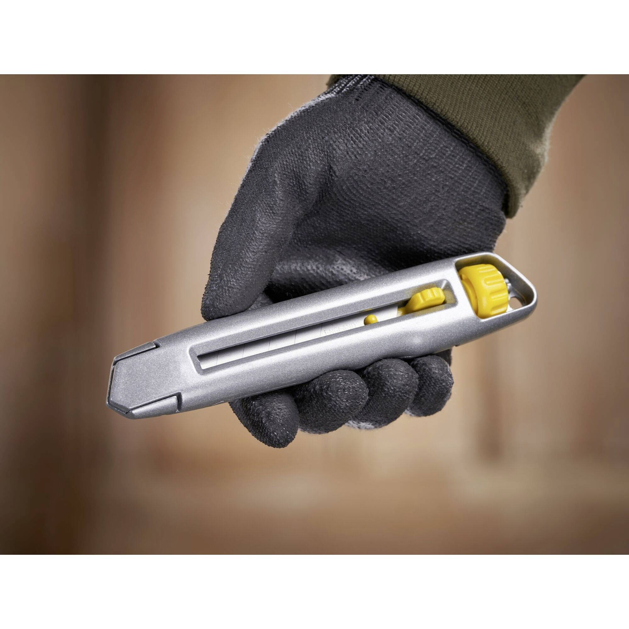 Eine behandschuhte Hand hält ein graues Universalmesser mit gelbem Schieber, bereit zum Schneiden.