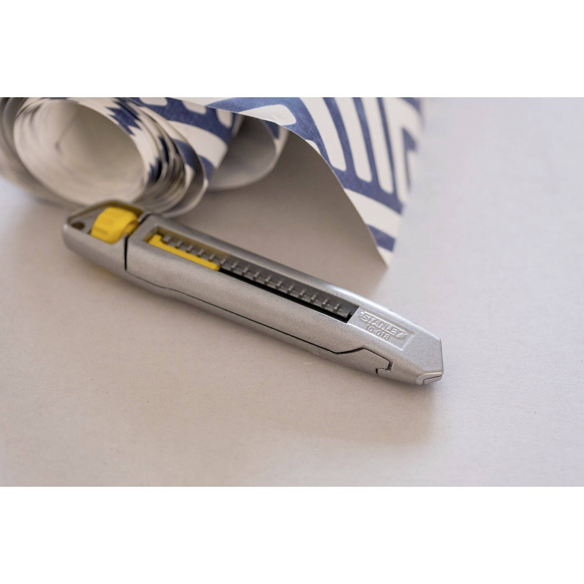 Ein silbernes Cuttermesser mit gelbem Schieber liegt auf einem weißen Tisch neben einer gerollten blauen und weißen Tapete.