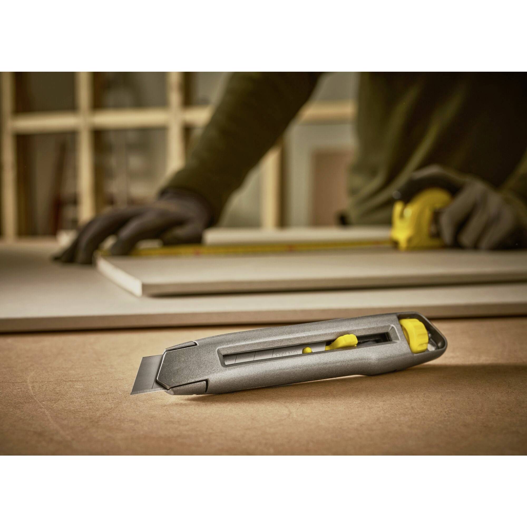 Ein Person misst ein Holzbrett mit einem Maßband, während ein Cuttermesser im Vordergrund liegt. Sie trägt Handschuhe.