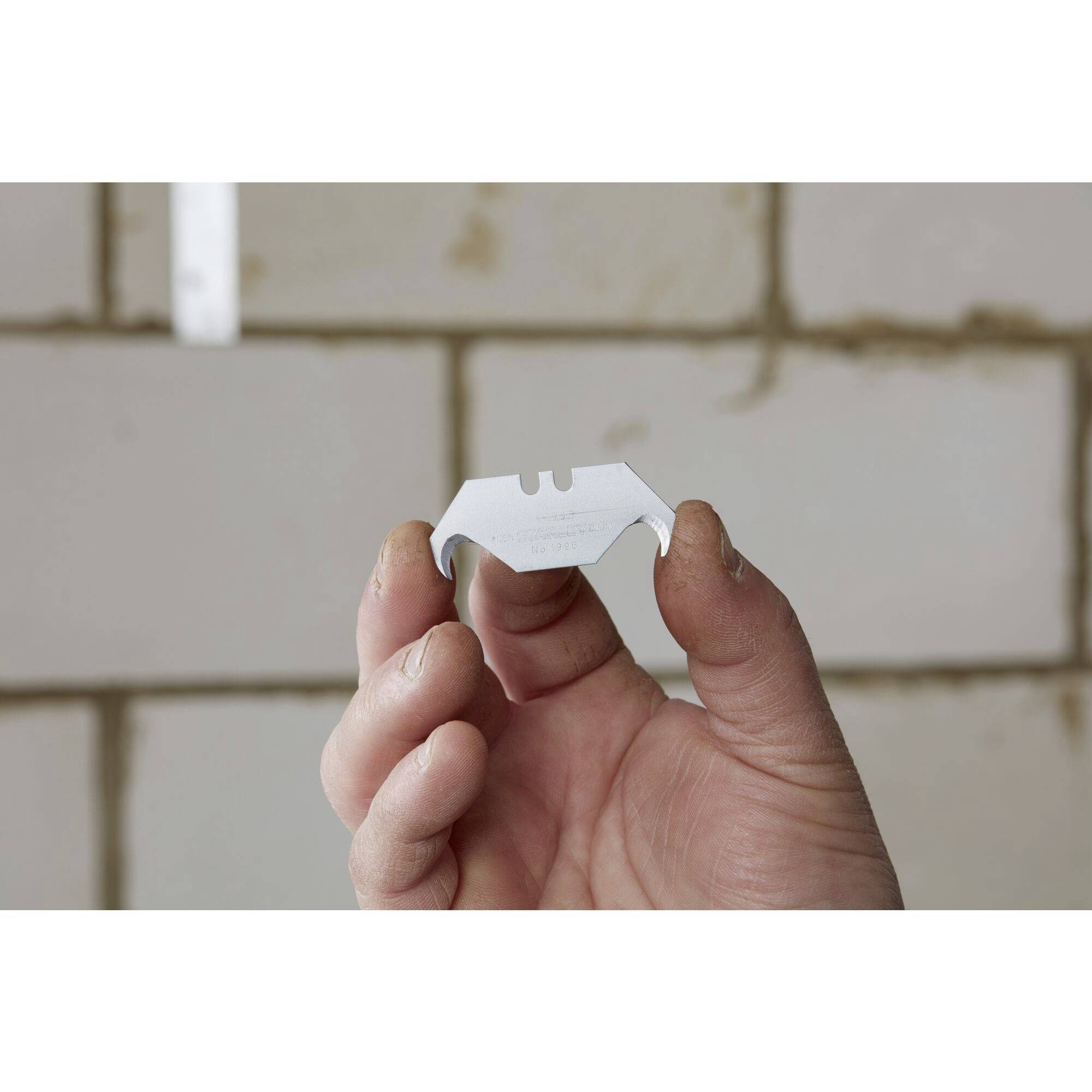 Eine Hand hält ein Rasierklingenblatt vor einer unscharfen Ziegelwand. Die Klinge wird vorsichtig mit den Fingerspitzen gehalten.