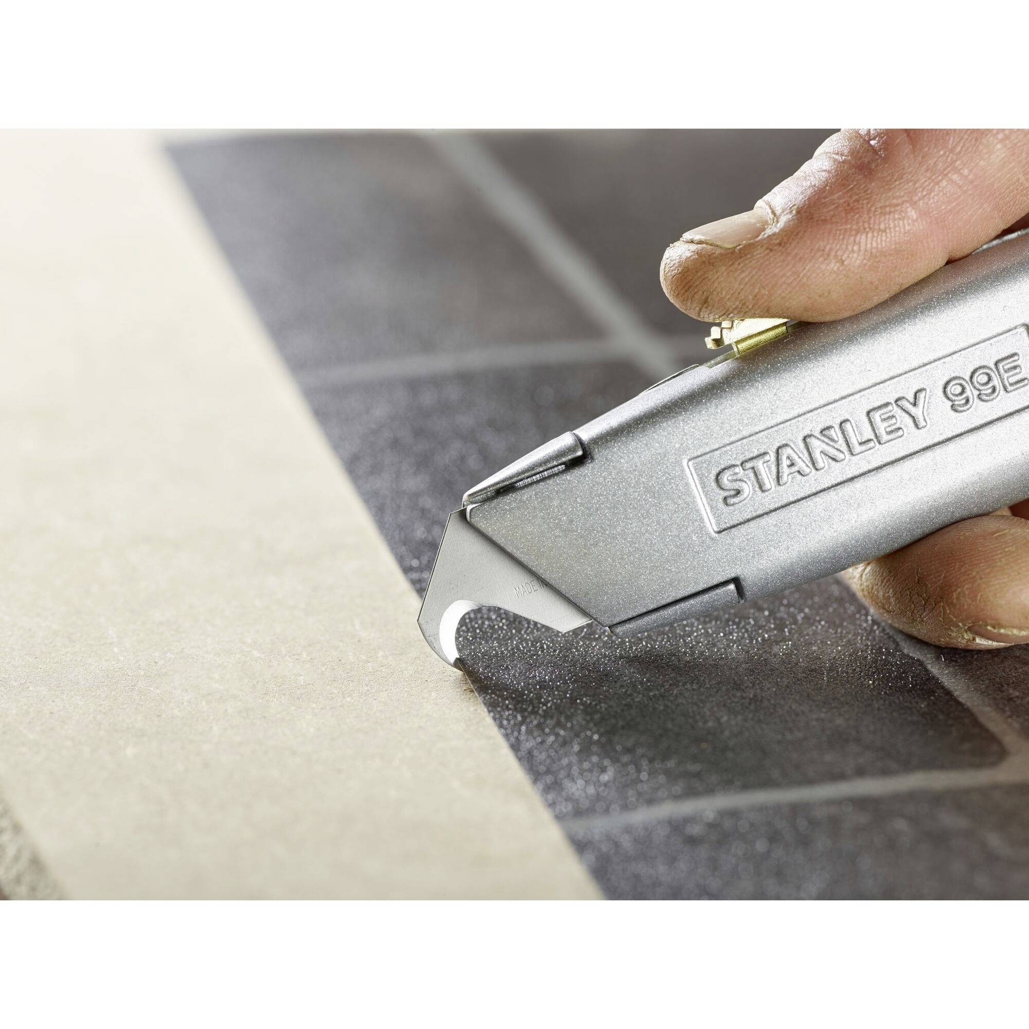 Eine Hand hält ein silbernes Stanley-99E-Universalmesser auf einer grauen Oberfläche, bereit zum Schneiden.