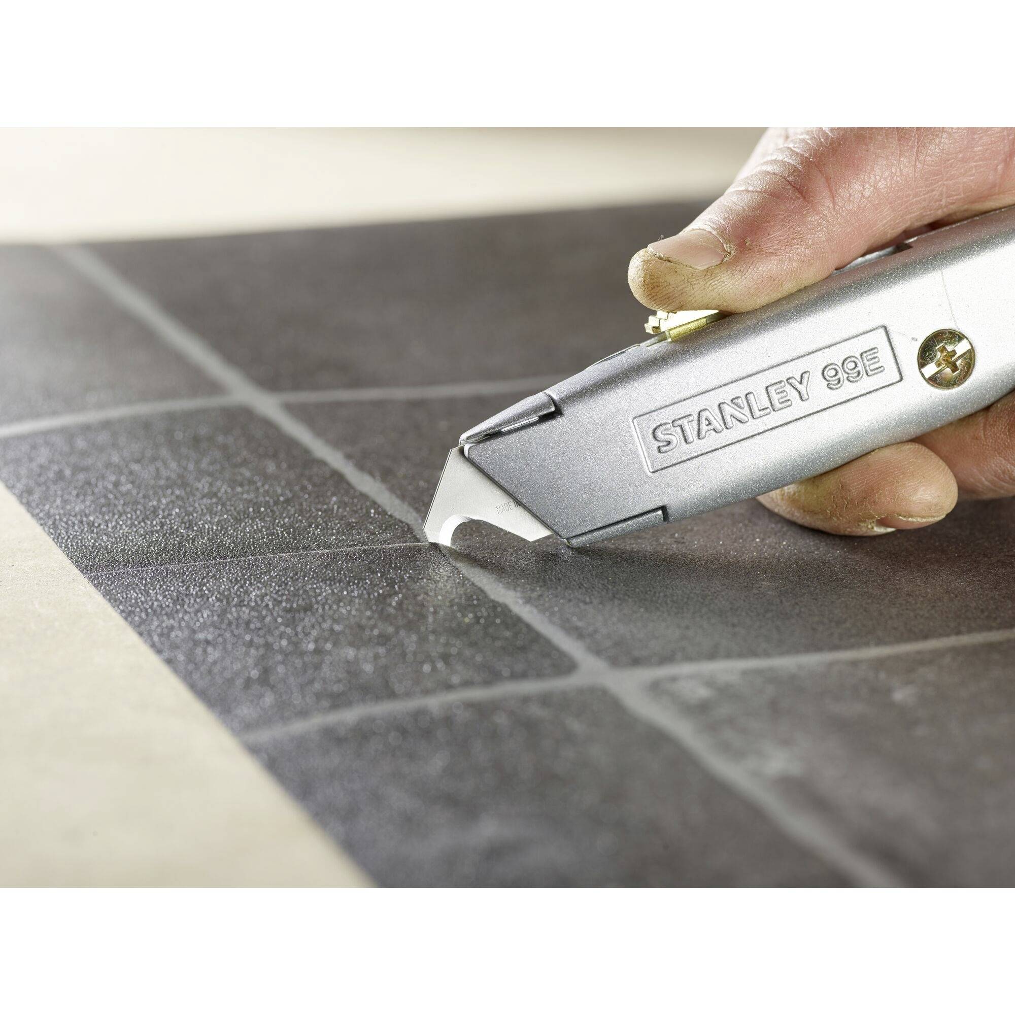 Eine Hand hält ein silberfarbenes Universalmesser mit dem Schriftzug 'Stanley 99E', schneidet präzise durch eine schwarze Fliesenoberfläche.