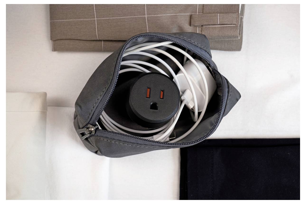 Offene schwarze Reißverschluss-Tasche mit aufgerolltem weißem Stromkabel und schwarzem Steckdosenadapter auf einem Bett oder Tisch.