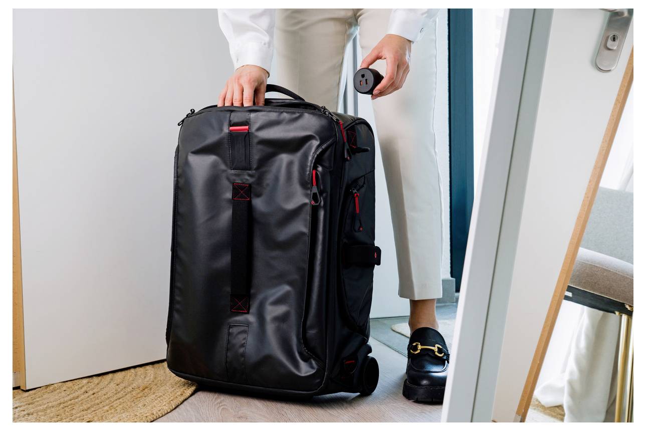 Eine Person packt einen großen schwarzen Rollkoffer in einer Türöffnung, hält den Griff und einen kleinen runden Gegenstand, was auf Reisen oder Abreise hindeutet.