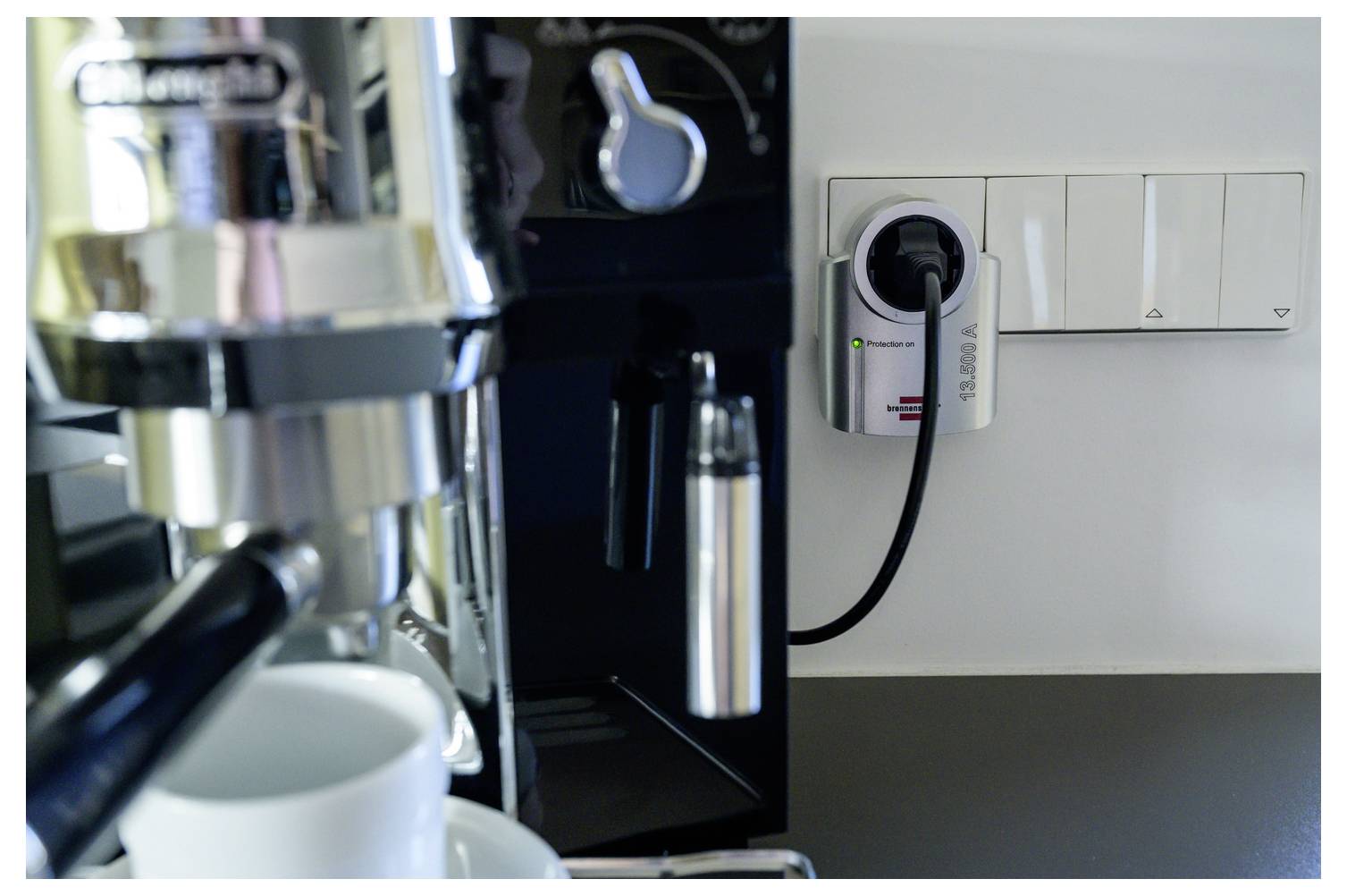 Nahaufnahme einer Kaffeemaschine, die in eine Wandsteckdose neben einem Lichtschalter-Panel eingesteckt ist, mit einer Tasse davor.