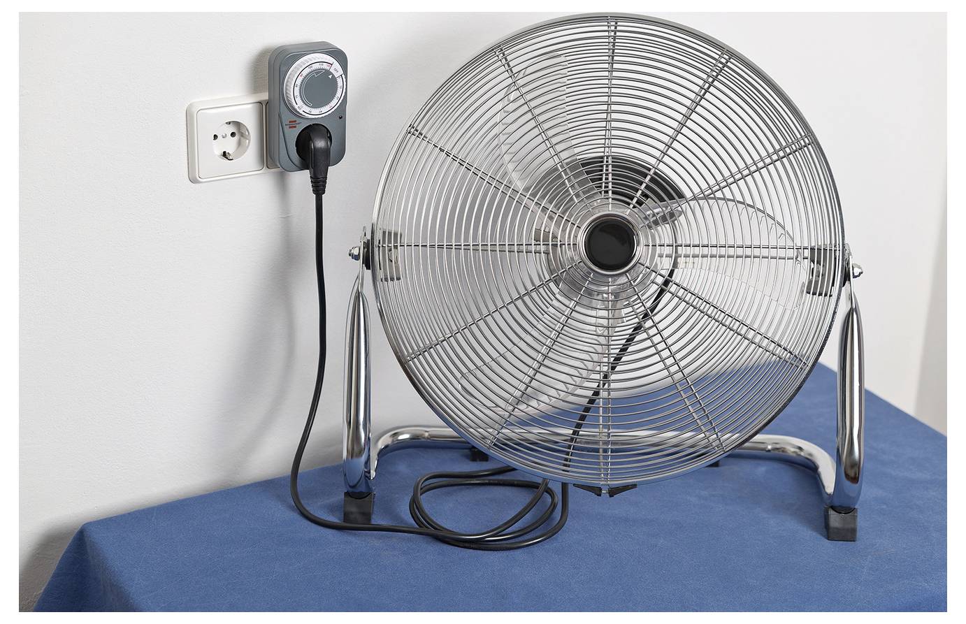 Großer elektrischer Bodenlüfter, der in eine Steckdose an einer blauen Oberfläche angeschlossen ist, mit einer Steuereinheit neben dem Stecker montiert.