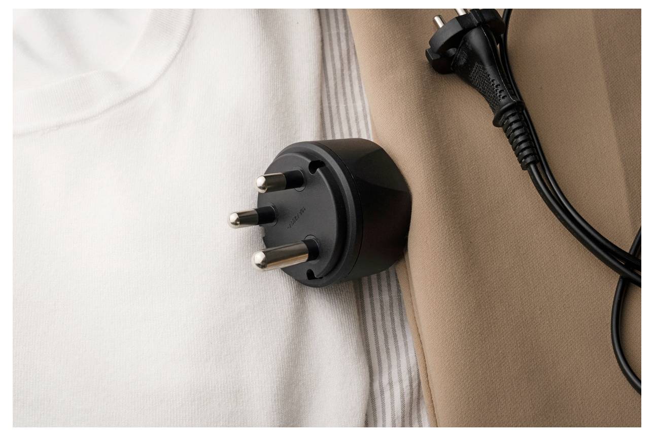 Nahaufnahme eines schwarzen dreipoligen Stromsteckers, der zwischen einer weißen und einer beige Oberfläche ruht, mit einem in der Nähe angeschlossenen Kabel.