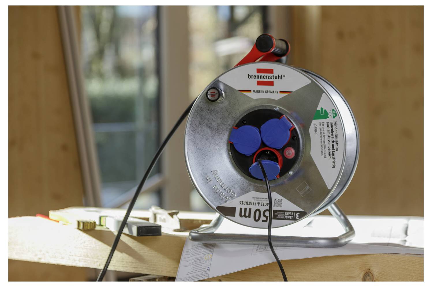 Eine Metallkabelrolle sitzt auf einer Holzoberfläche in einem sonnenbeschienenen Raum, mit zwei Kabeln, die in ihre blauen Buchsen eingesteckt sind.