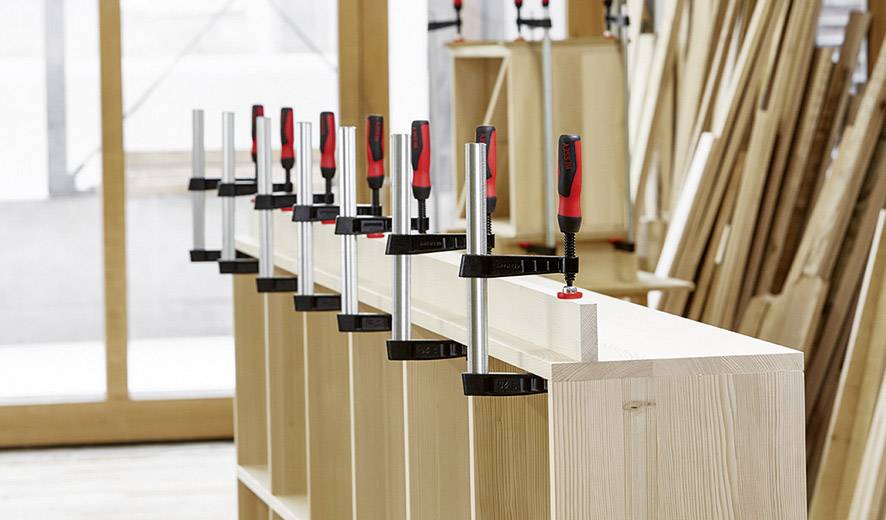 Mehrere Klemmen halten Holzbretter in einer Werkstatt zusammen, um ein größeres Möbelstück zu montieren. Holzmaterialien im Hintergrund.