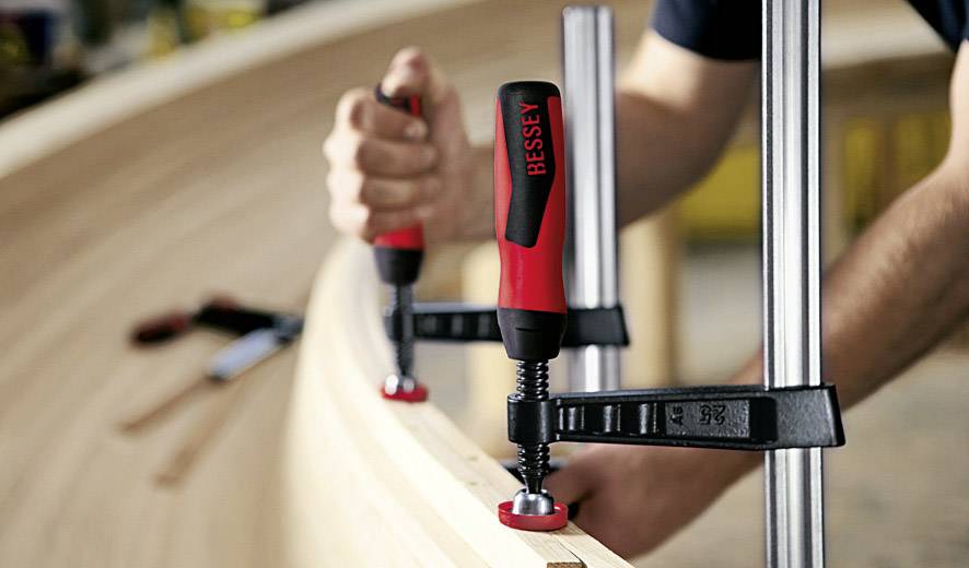 Eine Person verwendet rote Schraubzwingen, um ein hölzernes Bauteil in einer Werkstatt zu fixieren.