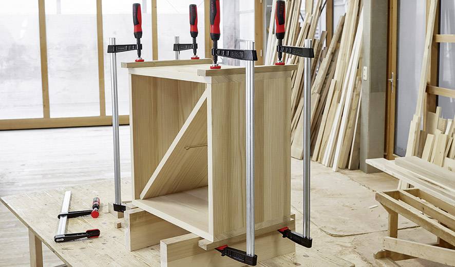 Ein Holzschrank wird mit Schraubzwingen auf einem Arbeitstisch zusammengebaut. Verschiedene Holzwerkzeuge befinden sich in der Nähe.