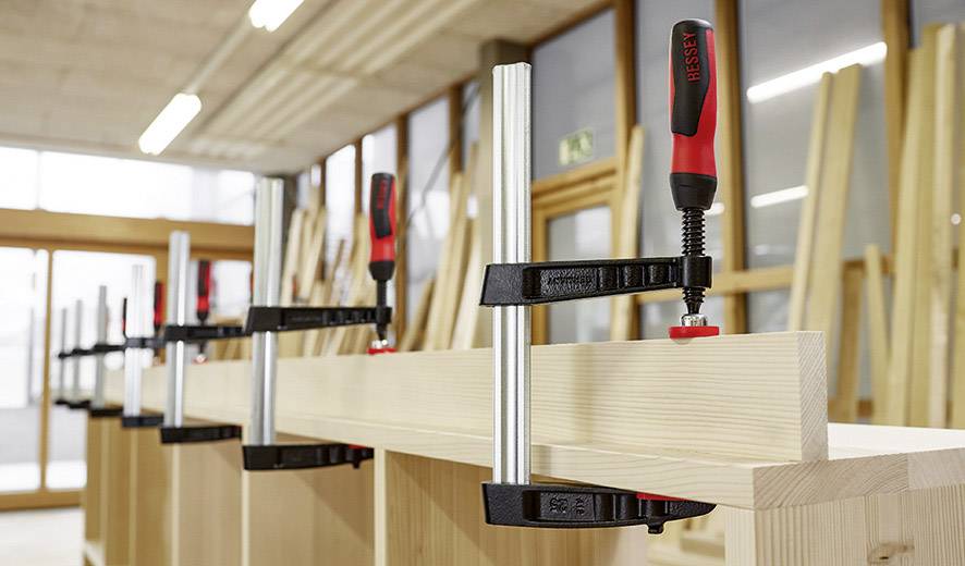 Holzbretter werden mit Zwingen auf einer Werkbank fixiert. Im Hintergrund sind weitere Holzbretter und eine Werkstatt zu sehen.