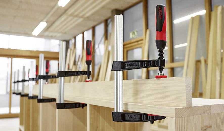 Werkbank mit Holzstücken, die mit mehreren Schraubzwingen gesichert sind. Hintergrund zeigt eine Werkstattumgebung.