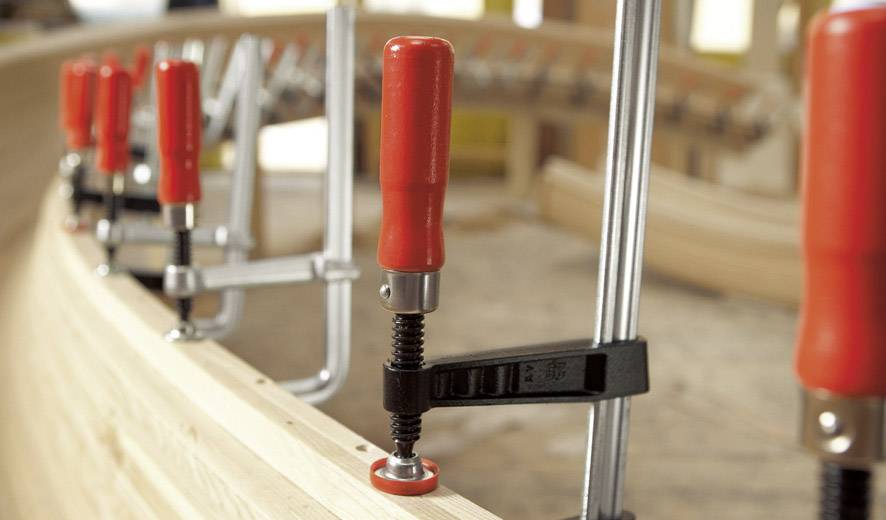 Holz wird mit mehreren roten Zwingen auf einer gebogenen Oberfläche fixiert, möglicherweise in einer Werkstatt.