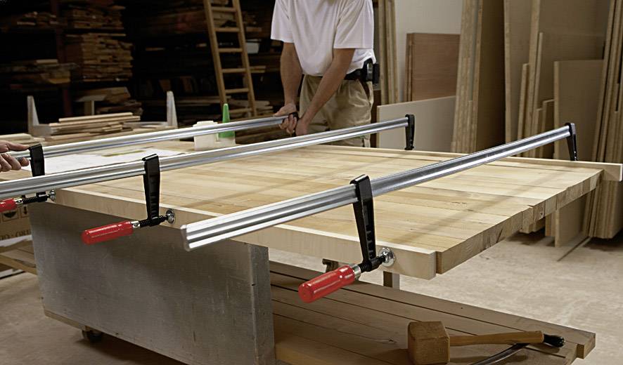 Zwei Personen arbeiten in einer Werkstatt mit Holzplatten, die auf einem Tisch mit großen Klemmen befestigt sind. Im Hintergrund sind gestapelte Holzvorräte sichtbar.