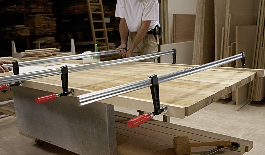 Zwei Personen spannen große Holzlatten mit Klemmen in einer Werkstatt ein. Die Arbeitsfläche ist mit Holzstücken und Werkzeugen gefüllt.