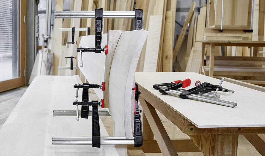 Holzwerkstatt mit Werkbank, auf der Holzplatten durch Schraubzwingen fixiert sind. Im Hintergrund sind Holzregale sichtbar.