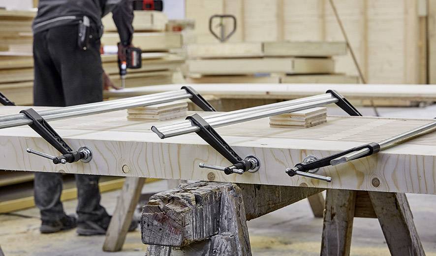 Eine Person arbeitet in einer Werkstatt, setzt Schraubzwingen auf verleimte Holzbretter, um sie zu fixieren. Im Hintergrund verschiedene Holzstücke.