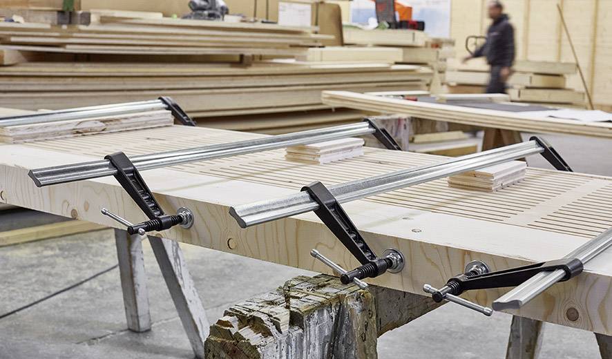 Holztischplatte wird in einer Werkstatt mit Metallzwingen fixiert. Im Hintergrund sind gestapelte Holzplatten und eine unscharfe Person zu sehen.