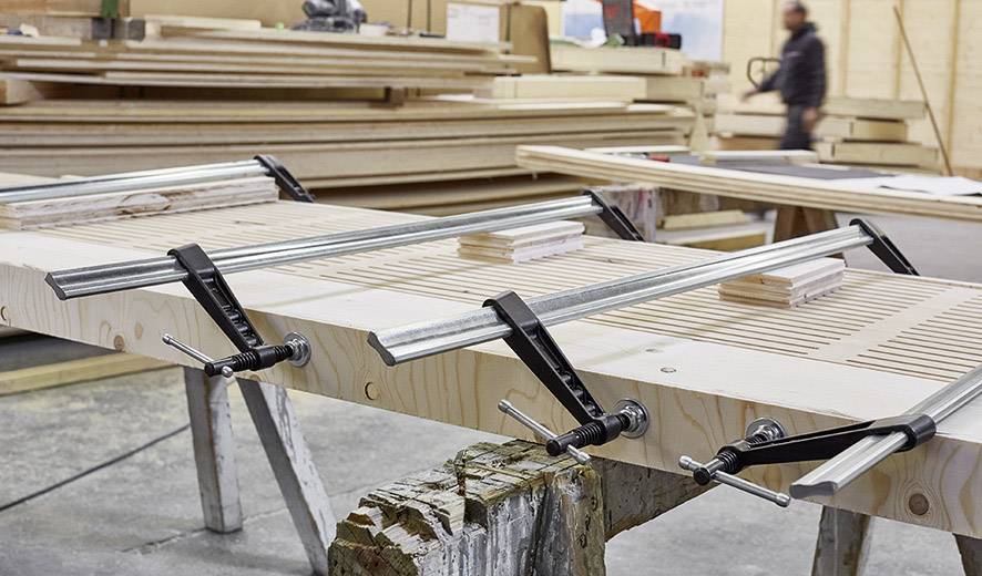 Holzplatte wird in einer Werkstatt mit Metallklemmen bearbeitet. Im Hintergrund stehen gestapelte Holzplatten und eine unscharfe Person.