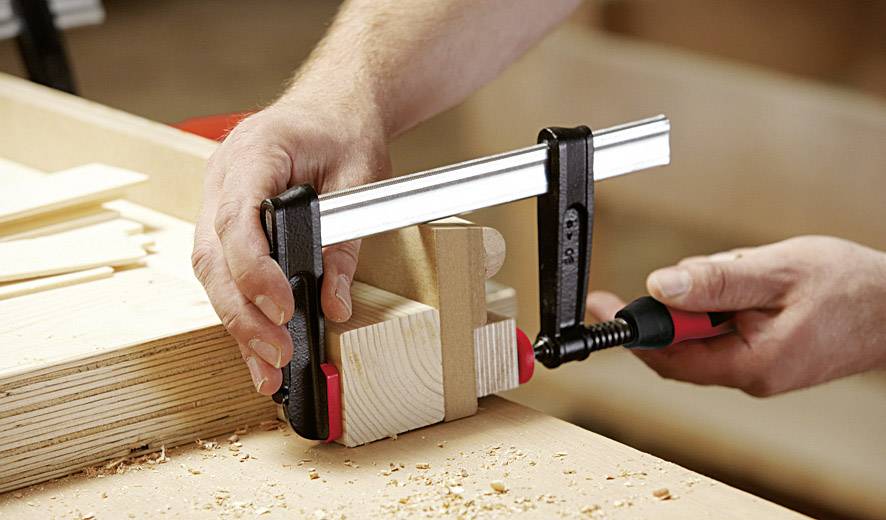 Eine Person klemmt ein Holzstück mithilfe einer Zwinge zum Sägen oder Schleifen auf einer Werkbank fest. Holzspäne sind sichtbar.