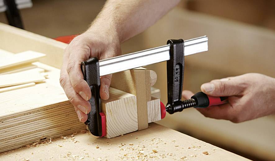Eine Person benutzt eine schwarze Zwinge, um Holzstücke auf einer Werkbank zu fixieren. Es sind Holzspäne sichtbar.