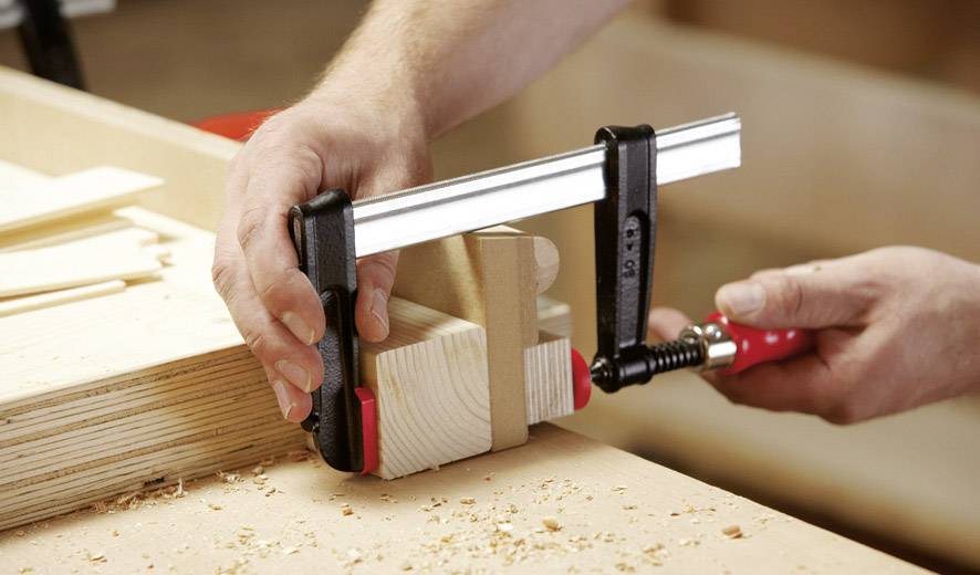 Eine Person befestigt Holzstücke mit einer Klemme auf einer Werkbank, um sie zu verleimen. Holzspäne liegen verstreut herum.