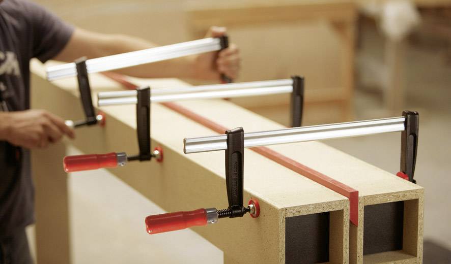 Eine Person, die Holzplatten mit langen Metallzwingen fixiert. Fokus liegt auf der Stabilisierung der Platten.