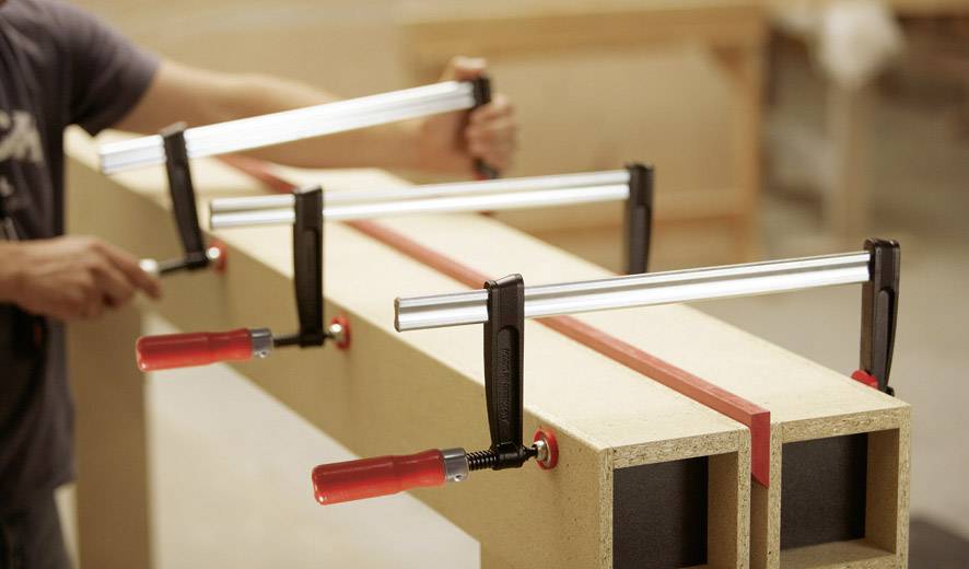 Eine Person befestigt Holzplatten mit vier großen Klemmen an einem Tisch. Die Klemmen haben rote Griffe und halten die Platten stabil.
