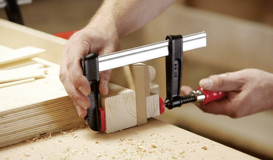 Eine Person benutzt eine Zwinge, um zwei Holzstücke auf einer Werkbank zu befestigen. Holzspäne sind auf der Oberfläche verteilt.