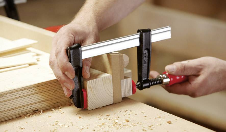 Eine Person klemmt zwei Holzstücke mit einer Zwinge zusammen. Im Hintergrund sind Werkstattutensilien und Holzspäne sichtbar.