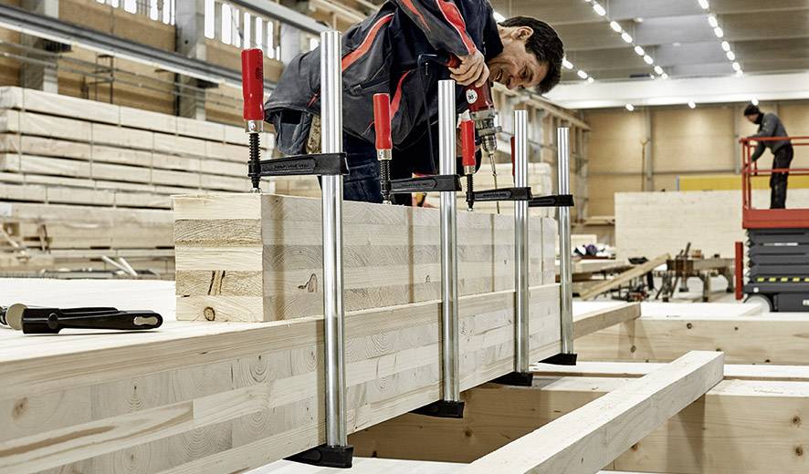 Ein Arbeiter in einer Werkstatt spannt Holzbohlen mit großen Klemmen fest. Die Werkstatt ist voller Holzwerkstoffe und Maschinen.