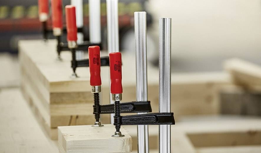 Mehrere rote Schraubzwingen befestigen Holzstücke an einer Werkbank in einer Werkstatt.