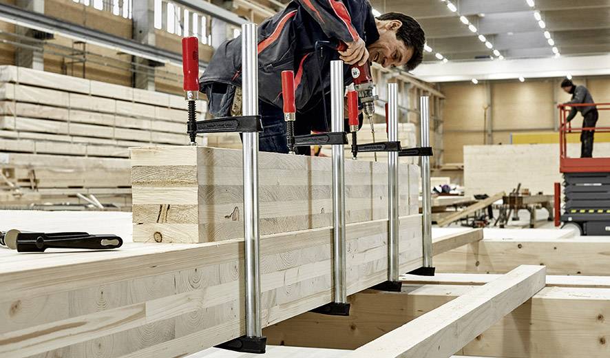 Eine Person bearbeitet Holz in einer Werkstatt, verwendet große Schraubzwingen, um Holzbalken zu fixieren. Im Hintergrund sind weitere Holzbalken und Bauarbeiter sichtbar, was eine aktive Arbeitsumgebung zeigt.