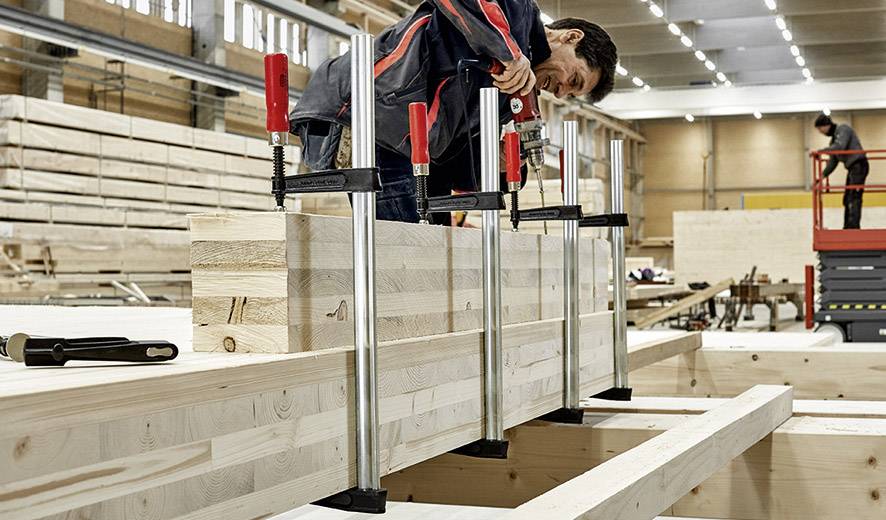 Ein Handwerker in einer Werkstatt bearbeitet mit Klemmen große Holzplatten. Im Hintergrund sind weitere Holzmaterialien und Klemmen zu sehen.