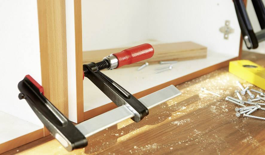 Holzwerkbank mit Schraubzwinge, die zwei Bretter verbindet. Auf der Oberfläche liegen Schrauben und ein gelber Wasserwaage.