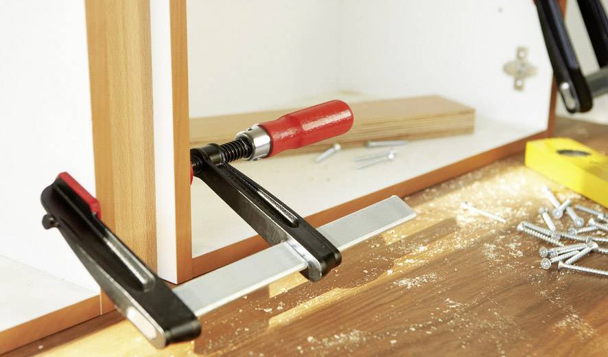 Holz-Werkstück an Schrank fixiert mit Schraubzwingen, umgeben von Schrauben und Holzspänen, helles Tageslicht auf Arbeitsfläche.