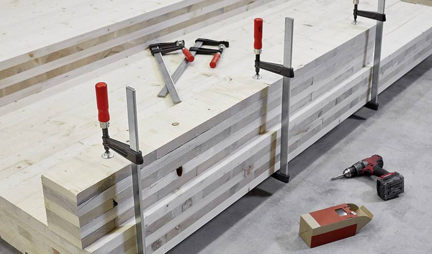 Stapeln von Holzplatten mit Schraubzwingen befestigt, rechts Bohrmaschine und Verpackung auf Betonboden.