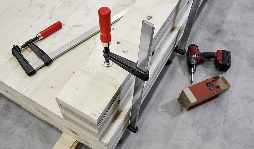 Holzplatte auf Werkbank mit Metallzwingen befestigt, daneben liegen eine elektrische Bohrmaschine und eine Packung Schrauben.