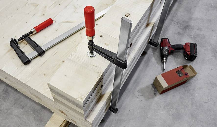 Ein Stapel Holz liegt auf dem Boden, mit einer Schraubzwinge befestigt. Daneben liegen eine Bohrmaschine und eine Packung Schrauben.