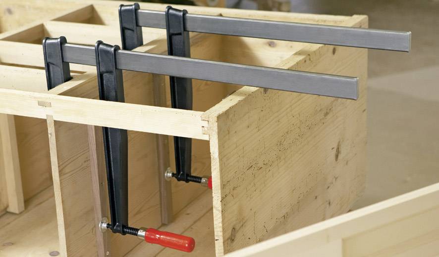 Zwei Holzbretter werden mit drei großen Schraubzwingen zusammengepresst, um eine stabile Ecke zu formen. Ideal für Holzarbeiten.