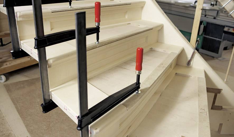 Eine Holztreppe im Bau, fixiert mit zwei großen roten Klemmen. Im Hintergrund sind Werkzeuge und Materialien einer Werkstatt erkennbar.