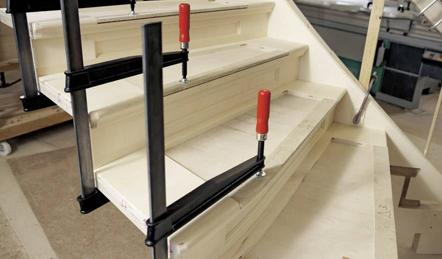 Eine halb fertige Holztreppe mit zwei Schraubzwingen, die die Stufen fixieren. Im Hintergrund sind Werkzeuge und Maschinen sichtbar.