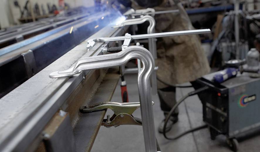 Ein Schweißer arbeitet in einer Werkstatt. Metallteile sind in einer Vorrichtung eingespannt. Funken fliegen beim Schweißen.