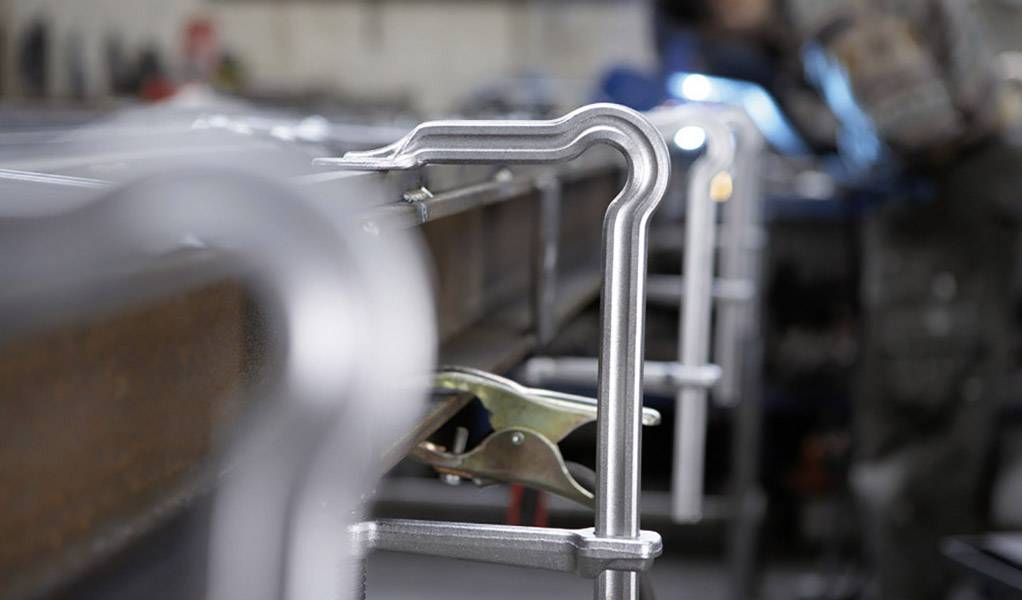 Eine metallene Schraubzwinge hält zwei Edelstahlplatten zusammen. Im Hintergrund verschwommen, eine Werkstatt mit Werkzeugen.
