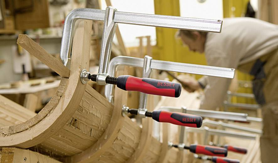 Eine Person arbeitet in einer Werkstatt und verwendet mehrere Schraubzwingen, um Holzstücke auf einer gewölbten Struktur zu fixieren.