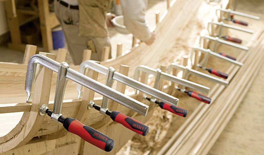 Mehrere Schraubzwingen halten Holzteile in Form eines Bootsrahmens zusammen. Eine Person arbeitet im Hintergrund am Holz.