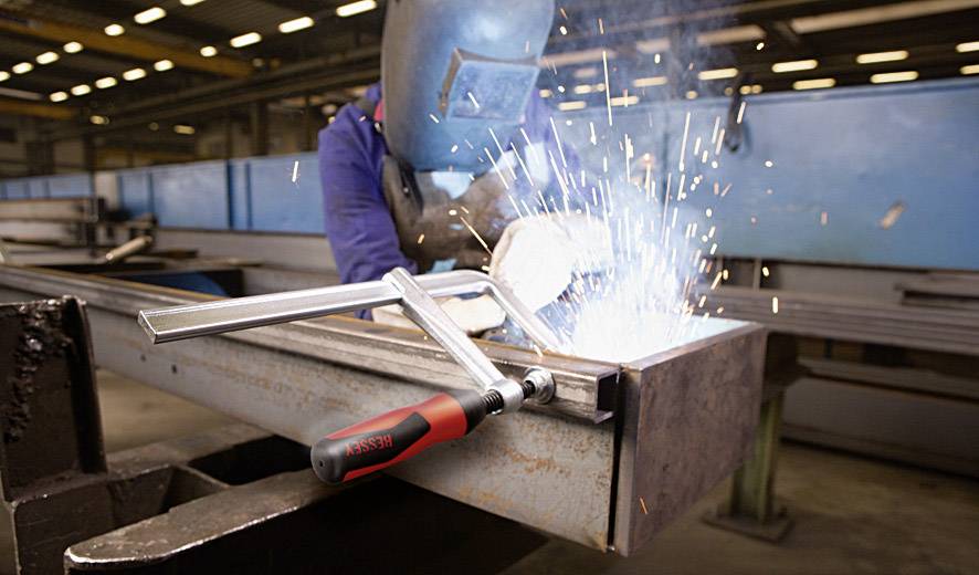 Ein Schweißer arbeitet in einer Fabrikhalle und erzeugt Funken. Im Vordergrund ist eine Klemme an einem Metallstück befestigt.