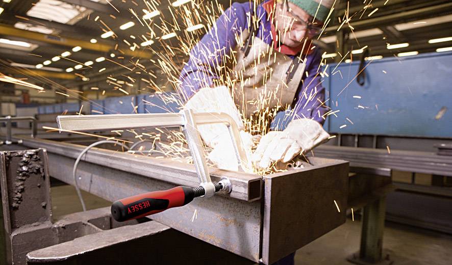 Ein Arbeiter schleift Metall in einer Werkstatt. Funken fliegen vom Werkstück. Der Arbeiter trägt Schutzkleidung, einschließlich Handschuhe und Helm.