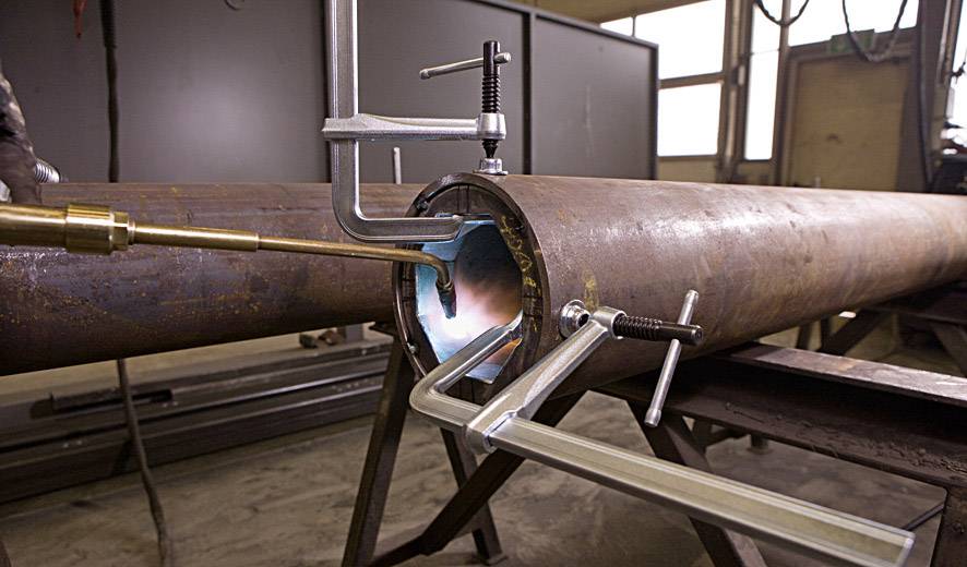 Eine Metallrohrverbindung wird in einer Werkstatt geschweißt. Verschiedene Klemmen halten die Rohre zusammen, während der Schweißvorgang stattfindet.
